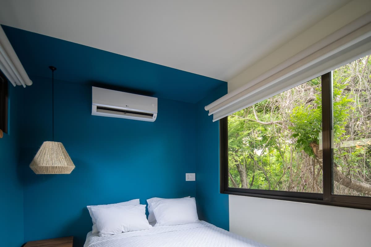 A bedroom with a bed, a bedside table, an air conditioning unit, and a window with a view of greenery.