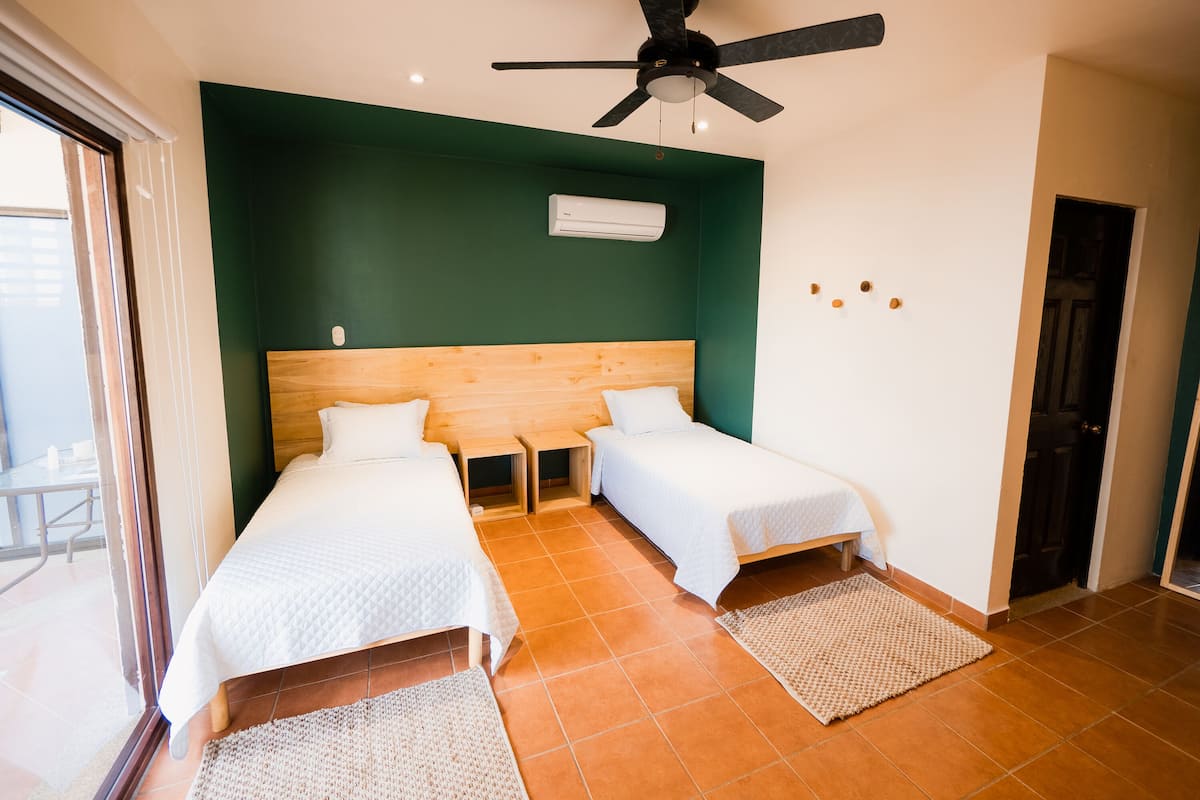 A room with two single beds, a ceiling fan, an air conditioning unit, and a view of the outside.
