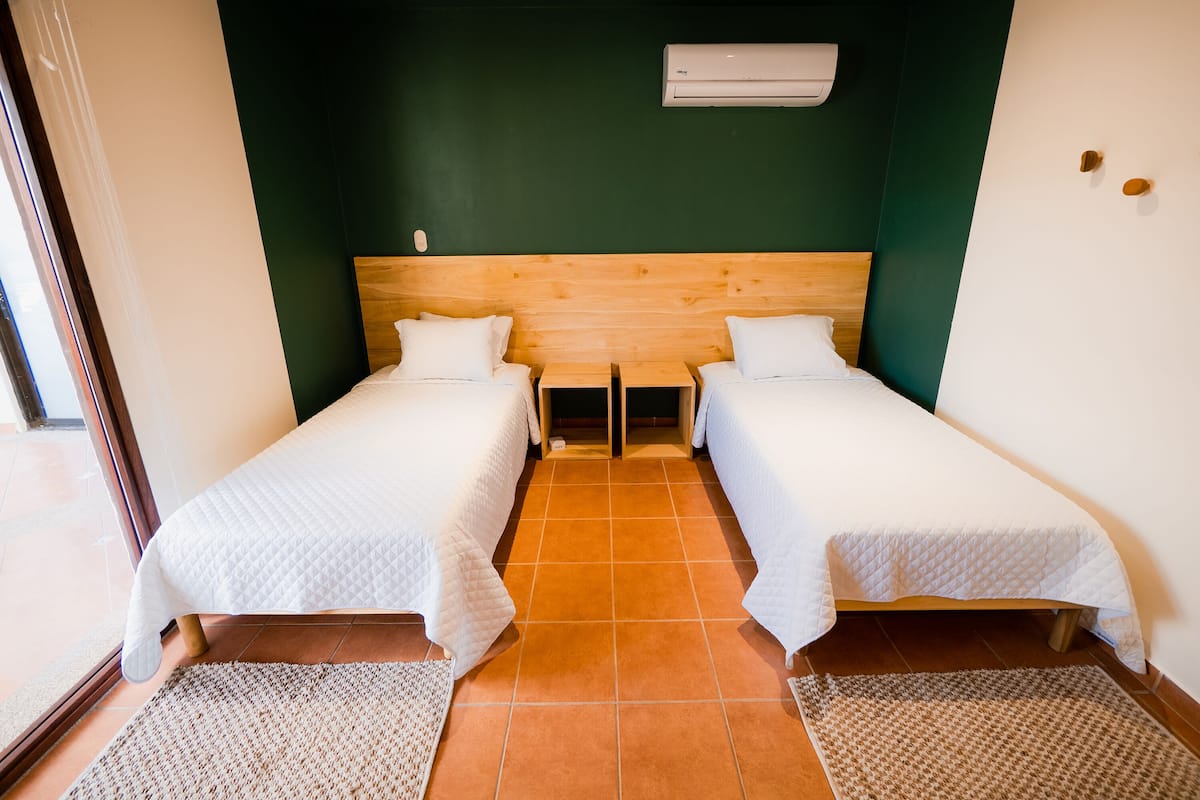 A room with two single beds, a wooden headboard, an air conditioning unit, and a tiled floor.