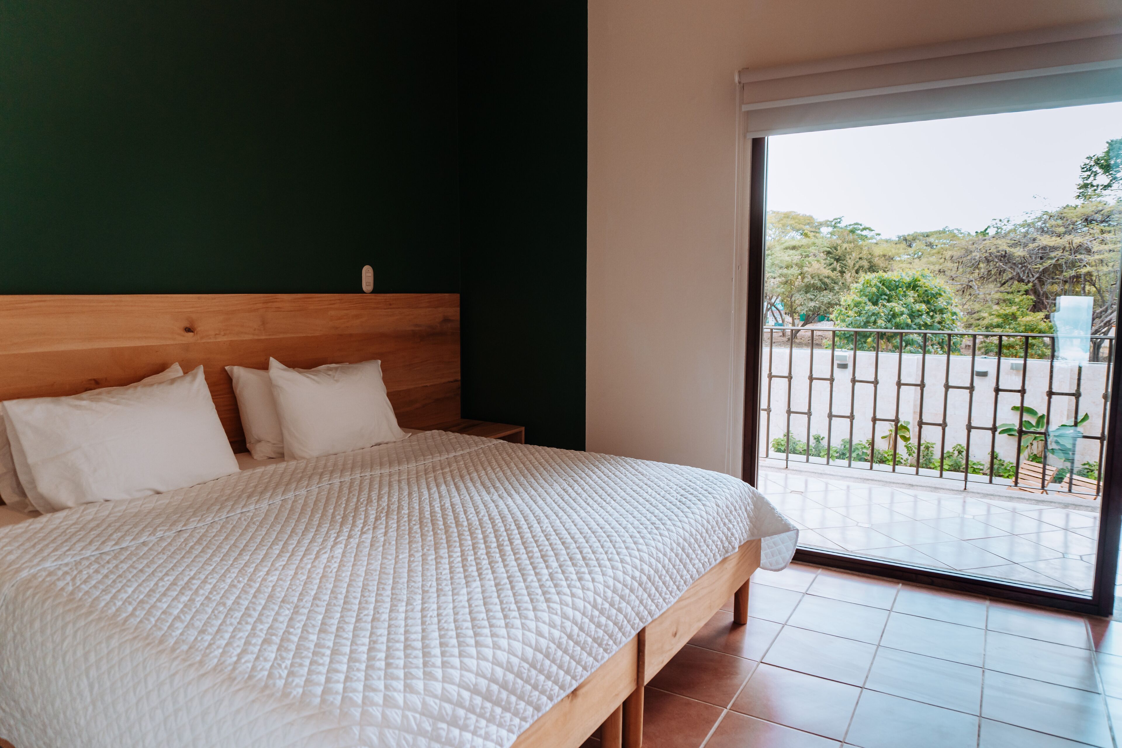 A bedroom with a wooden headboard, a white quilted bedspread, and a view of a balcony with a metal railing and greenery.