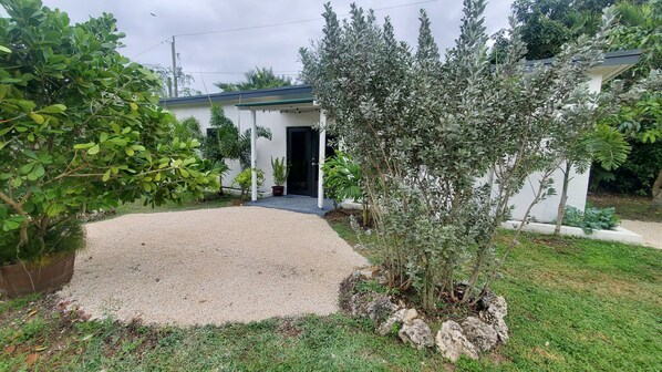 A residential property with a gravel driveway, a white building with a dark entrance, and various greenery including a large tree and shrubs. - Stylish cozy modern open concept guest house (Homestead)