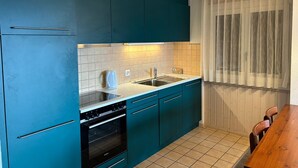 A compact kitchen with teal cabinets, a white countertop, a built-in oven, and a window with sheer curtains.