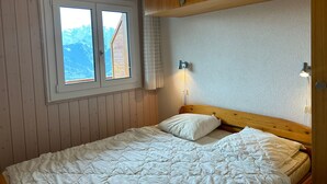 A bedroom with a wooden headboard, a white quilted bedspread, a window with a view of mountains, and a wall-mounted light fixture.