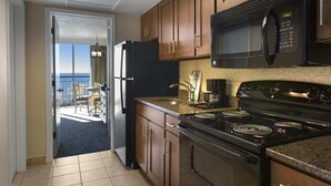 Cucina con mobili in legno, forno a microonde nero, piano cottura e frigorifero. Si vede l'oceano attraverso una porta.