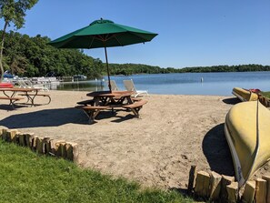 A lakeside picnic area with wooden benches, a green umbrella, and a canoe. - Swan Lake Resort & Campground - Lakeside Cabin #1 (Fergus Falls)