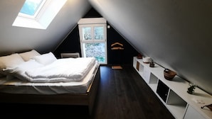 Quarto com teto inclinado, cama com roupa de cama branca, janela e cómoda branca com gavetas.
