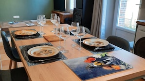 Uma sala de jantar com uma longa mesa de madeira posta para refeição, cadeiras ao redor e uma televisão ao fundo.