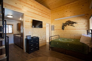 A wooden cabin interior with a bed, dresser, TV, and a mounted plant.