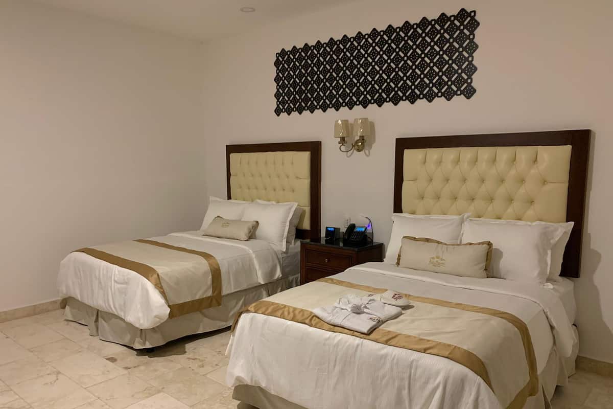 A hotel room with two beds, each with a tufted headboard and white bedding with gold accents. There is a decorative wall panel above the beds and a nightstand with a phone and lamp between them.