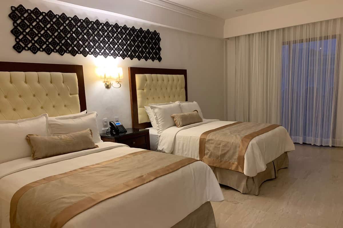 A hotel room with two beds, a decorative wall panel, a nightstand with a phone, and a large window with curtains.
