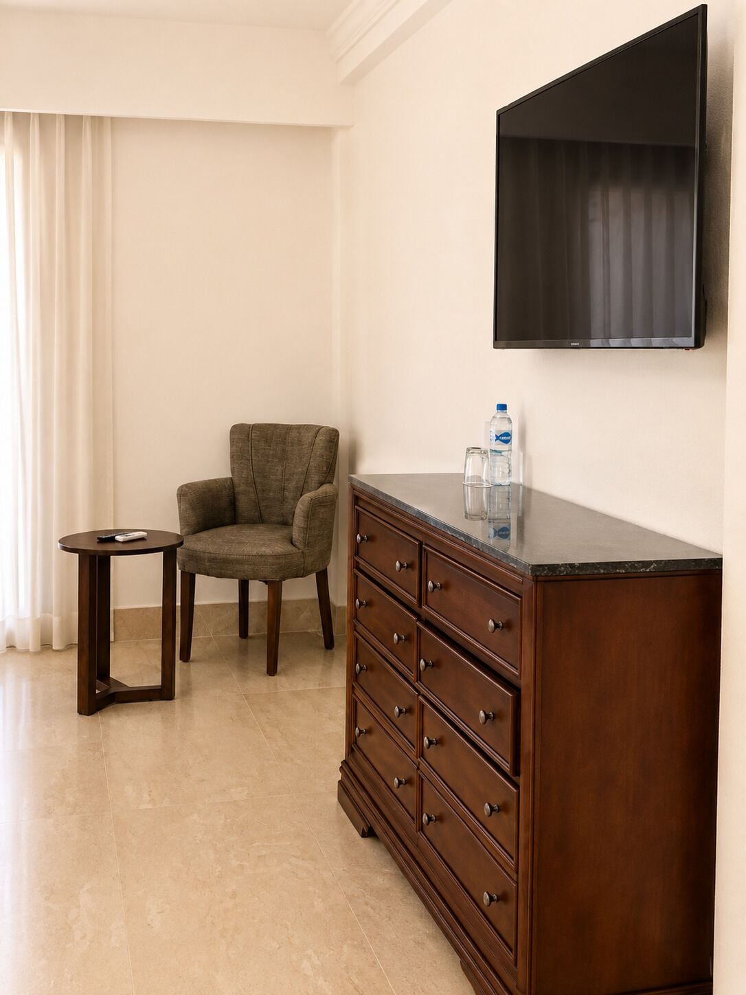 Une pièce comprenant une commode en bois, une chaise, une petite table et un téléviseur à écran plat fixé au mur.