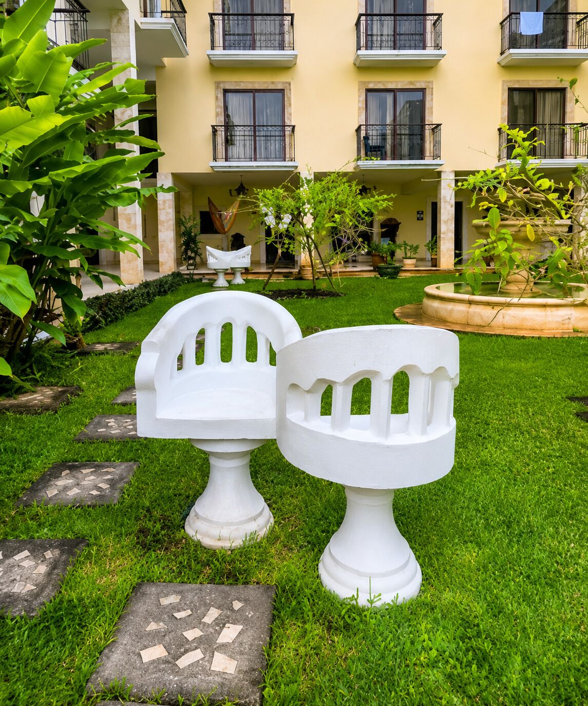 Un patio con dos bancos blancos de jardín, una fuente y un edificio con balcones al fondo.