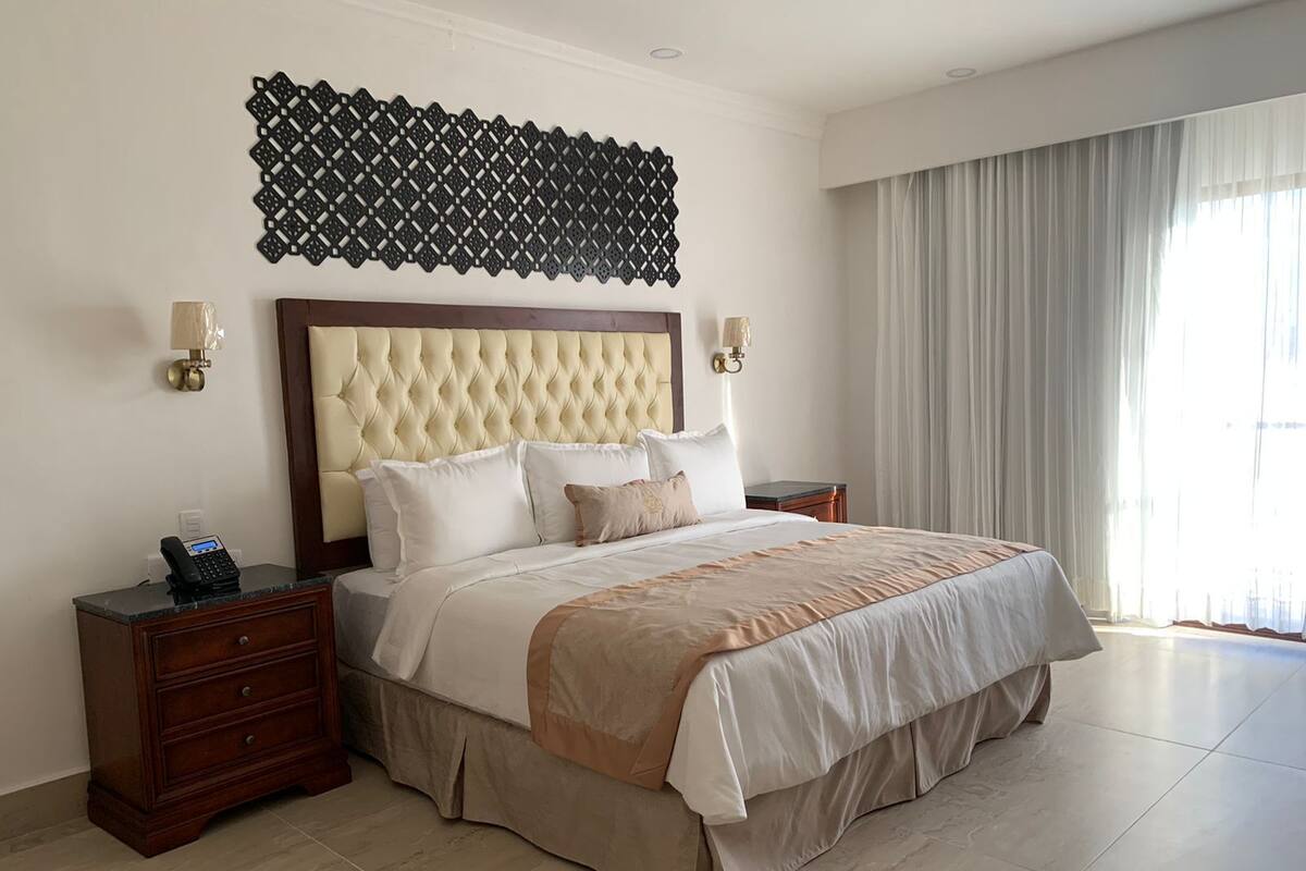 A large bed with a tufted headboard, two bedside tables with lamps, a decorative wall piece, and a large window with curtains.
