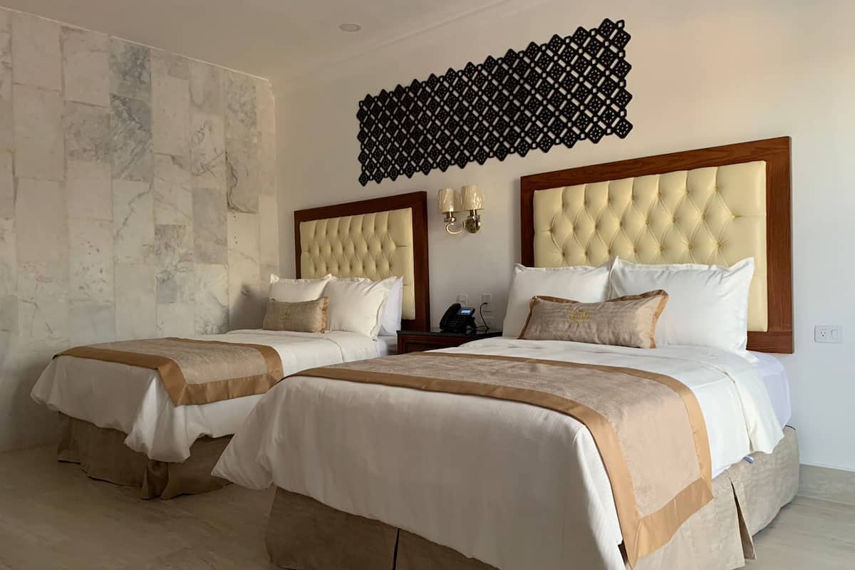 A hotel room with two beds, each with a tufted headboard and beige bedding. There is a decorative wall panel above the beds and a wall-mounted light fixture.