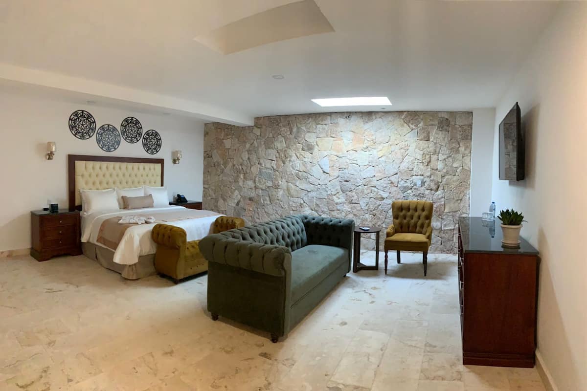 A bedroom with a bed, a green sofa, a yellow armchair, a small table, and a stone wall.