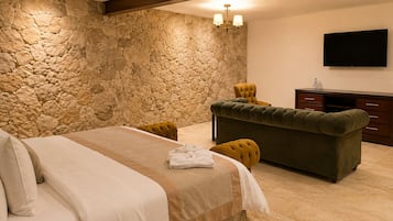 Un dormitorio con pared de piedra, cama con cubrecama beige y marrón, sofá verde y televisor de pantalla plana en la pared.
