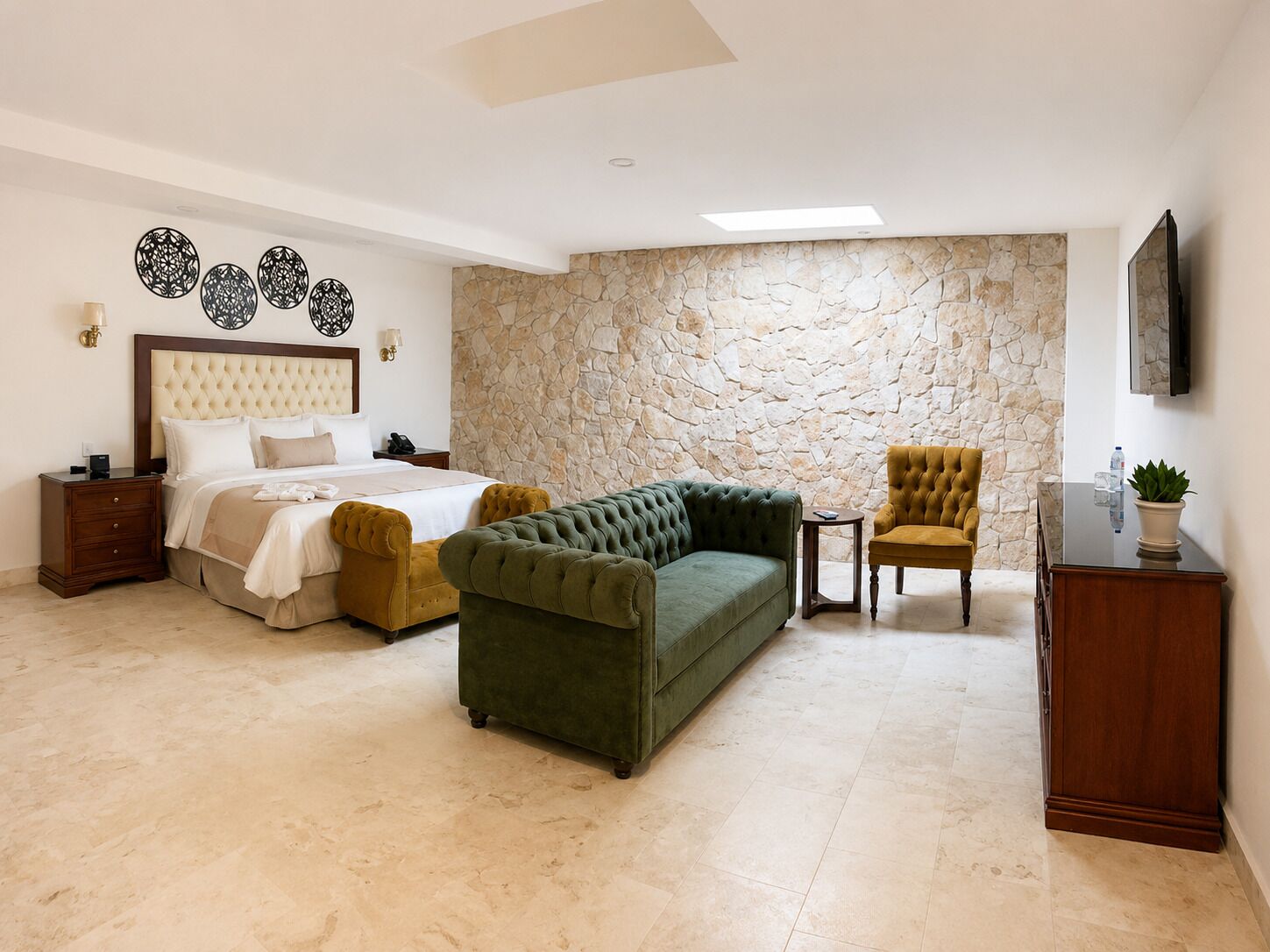 A bedroom with a bed, a green sofa, a yellow armchair, a stone wall, and a TV mounted on the wall.