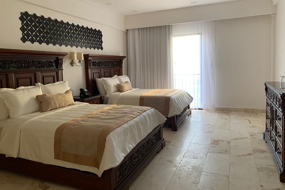 A hotel room with two beds, a wooden headboard, a decorative wall piece, a large window with curtains, and a dresser.