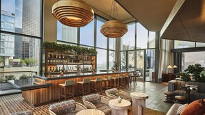 A modern bar area with a wooden bar counter, seating, and large windows offering city views. - Conrad Los Angeles (Los Angeles)