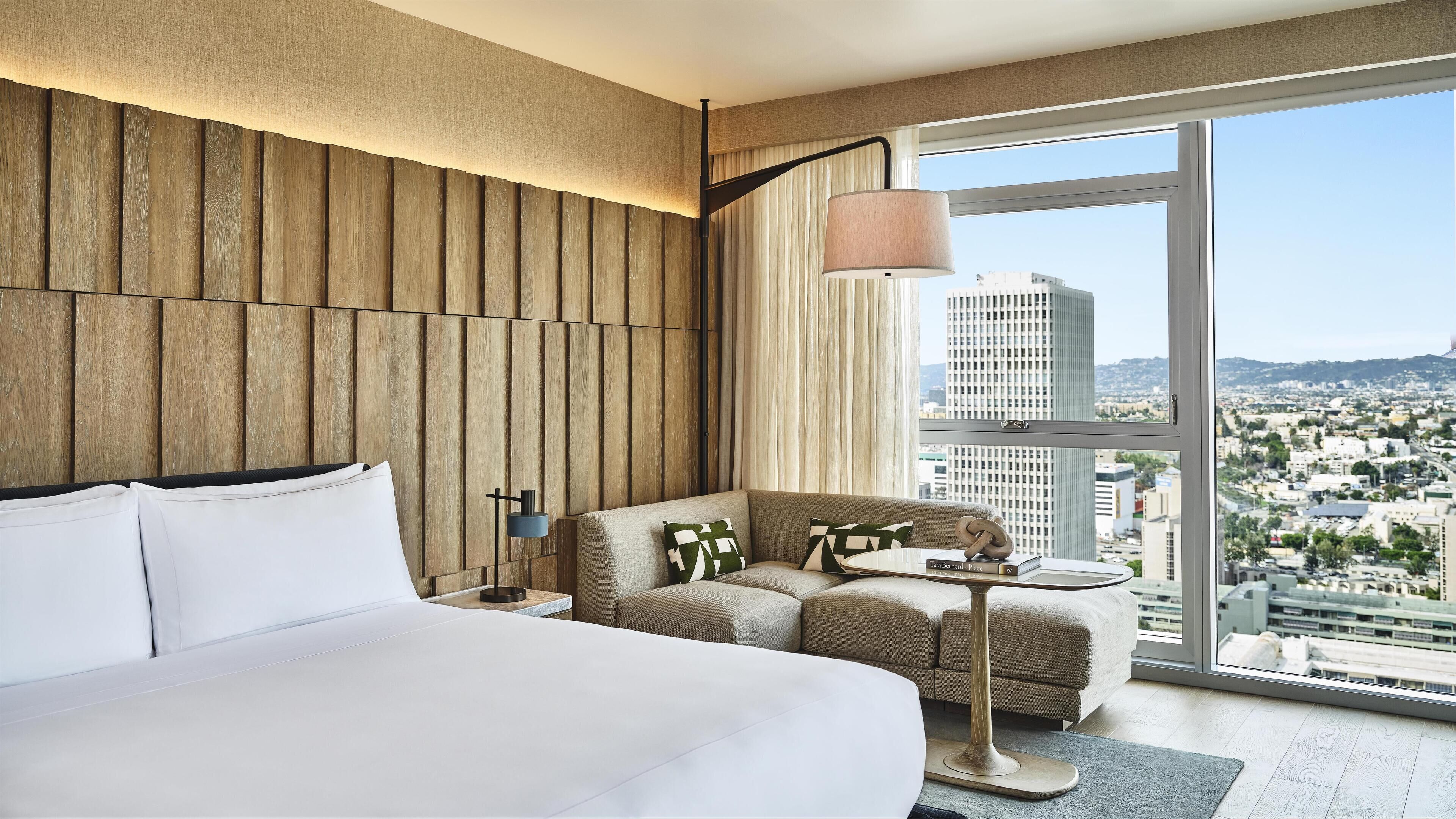Habitación de hotel moderna con una cama grande, un sofá, una mesita y vistas a la ciudad a través de amplios ventanales.