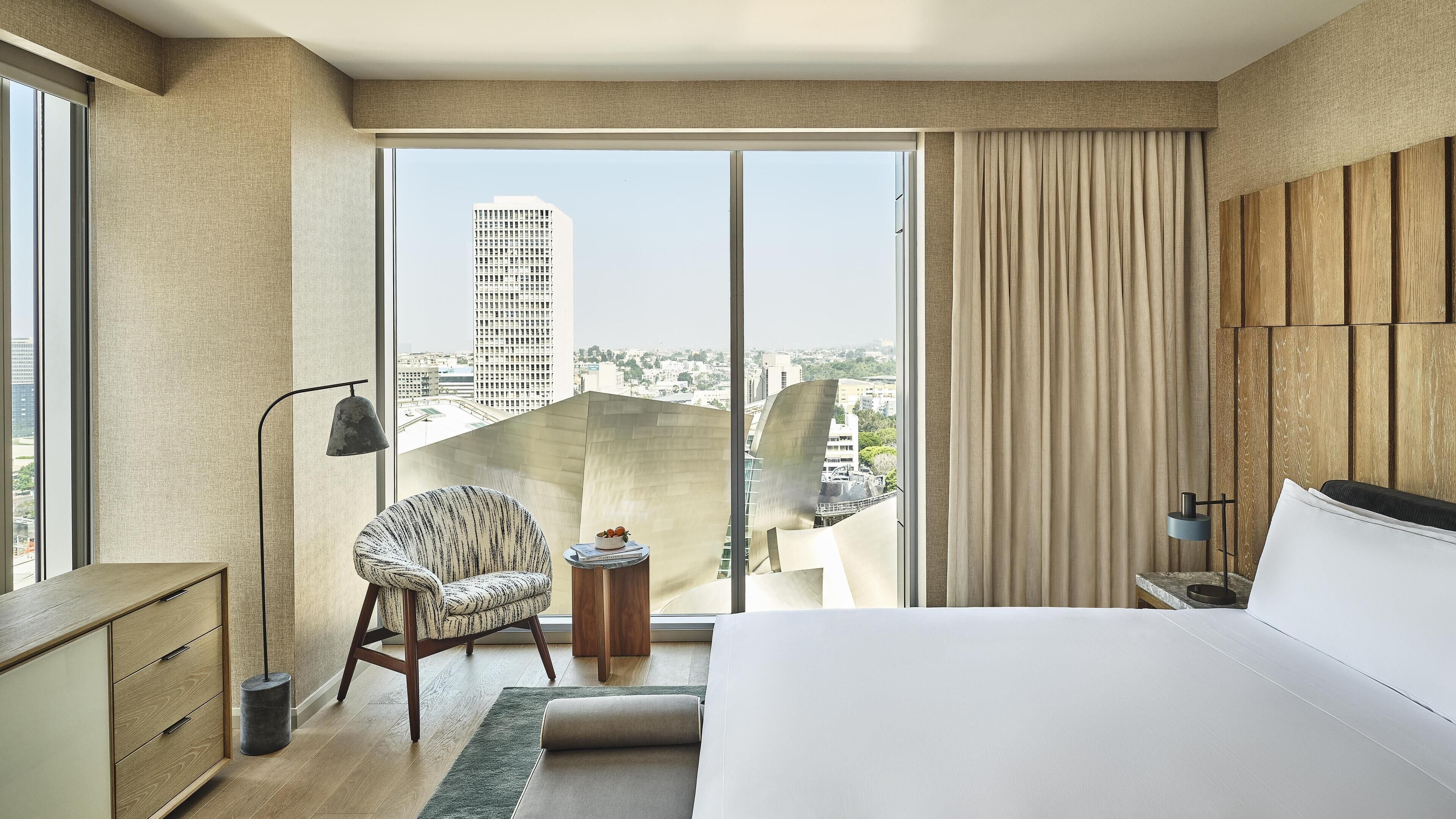 A modern hotel room with a large bed, a chair, a small table with a bowl of fruit, and a view of a cityscape through a sliding glass door.