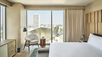 A modern hotel room with a large bed, a chair, a small table with a bowl of fruit, and a view of a cityscape through a sliding glass door.