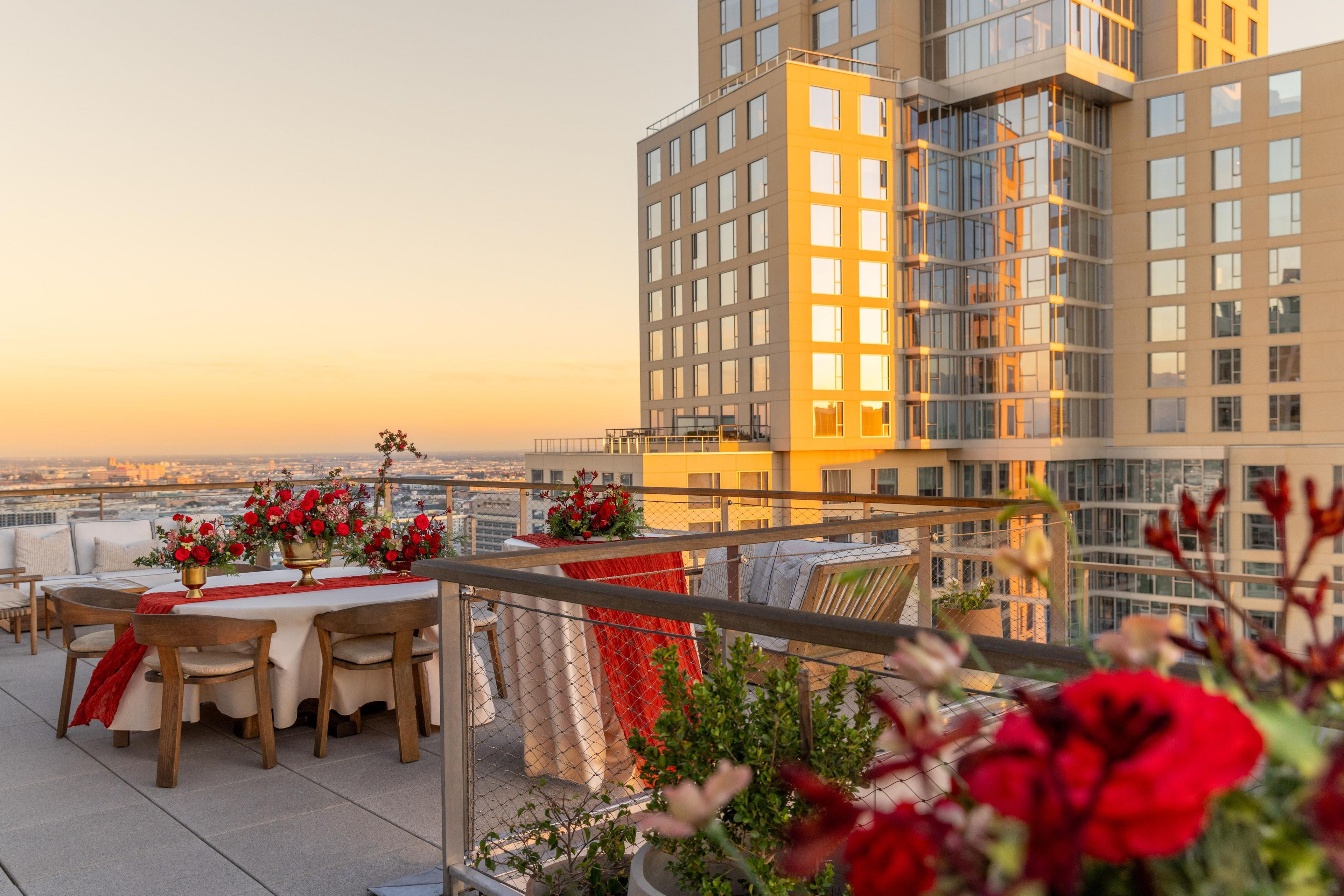 A rooftop terrace with a dining table set for two, surrounded by red flowers and a view of the cityscape.