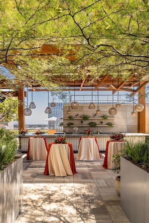 An outdoor dining area with tables covered in white and red tablecloths, hanging pendant lights, and potted plants. - Conrad Los Angeles (Los Angeles)