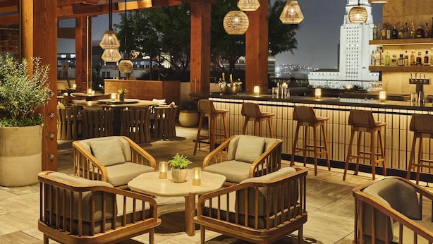 A rooftop bar with wooden furniture, hanging pendant lights, and a view of a city skyline. - Conrad Los Angeles (Los Angeles)