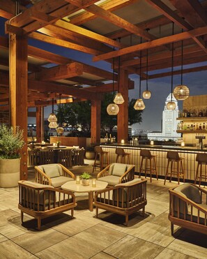 A rooftop bar with wooden furniture, hanging pendant lights, and a view of a city skyline. - Conrad Los Angeles (Los Angeles)