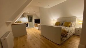 A bedroom with a bed, bedside tables, and a nightstand. There is a small kitchenette area with a sink and a stove. The room has wooden flooring and white walls.