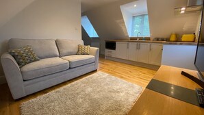 A modern loft apartment with a grey sofa, wooden flooring, a kitchen with white cabinets, and a skylight.