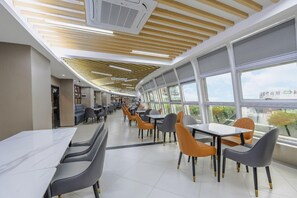 Ein moderner Essbereich mit langen Tischen und Stühlen, großen Fenstern und einer Decke mit Holzleisten.