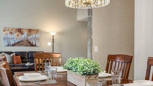 A dining area with a wooden table set for four, surrounded by wooden chairs. A chandelier hangs above, and a painting is visible on the wall.