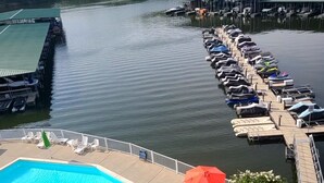 Ein Schwimmbecken mit Liegestühlen und Sonnenschirmen, eine Anlegestelle mit Booten und ein bewaldeter Uferstreifen.