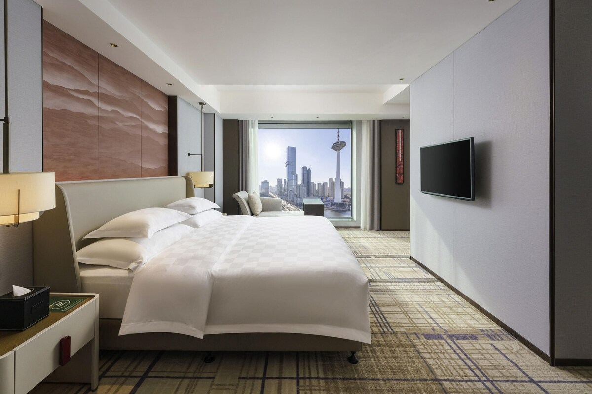 A hotel room with a large bed, a TV, a nightstand, and a view of a cityscape.