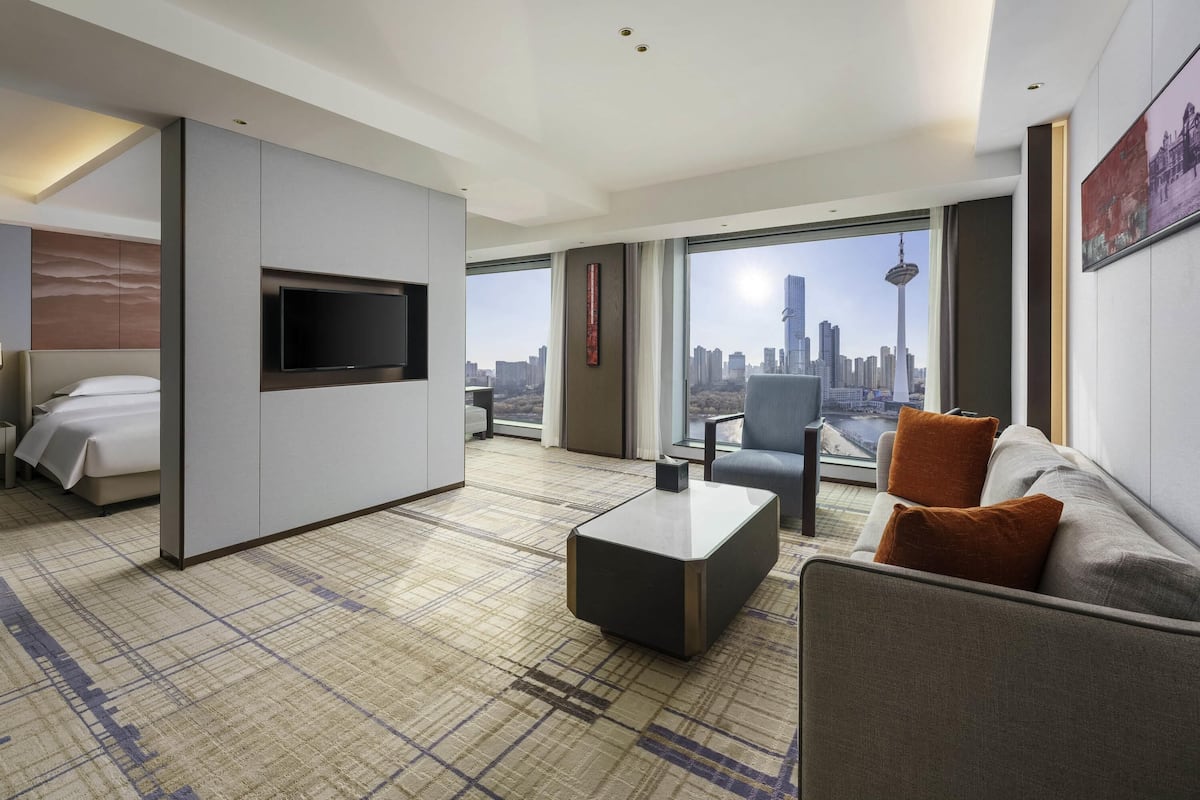 A modern hotel room with a bed, a sofa, a TV, a coffee table, and a large window offering a city view.