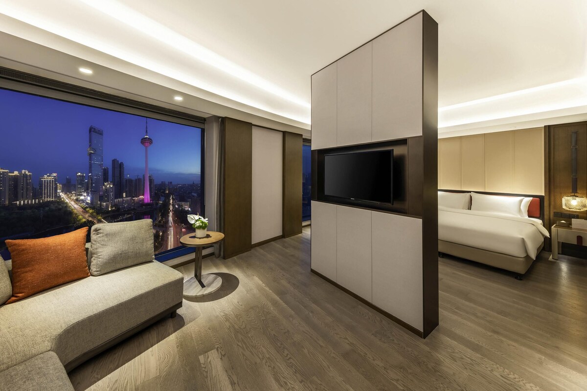 A modern hotel room with a large TV, a sofa, a bed, and a view of a cityscape at dusk.