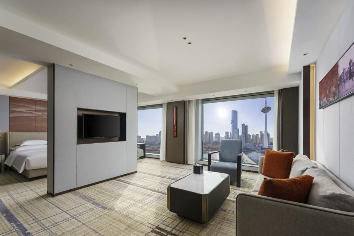 A modern hotel room with a large bed, a flat-screen TV, a sofa, a coffee table, and a city view.