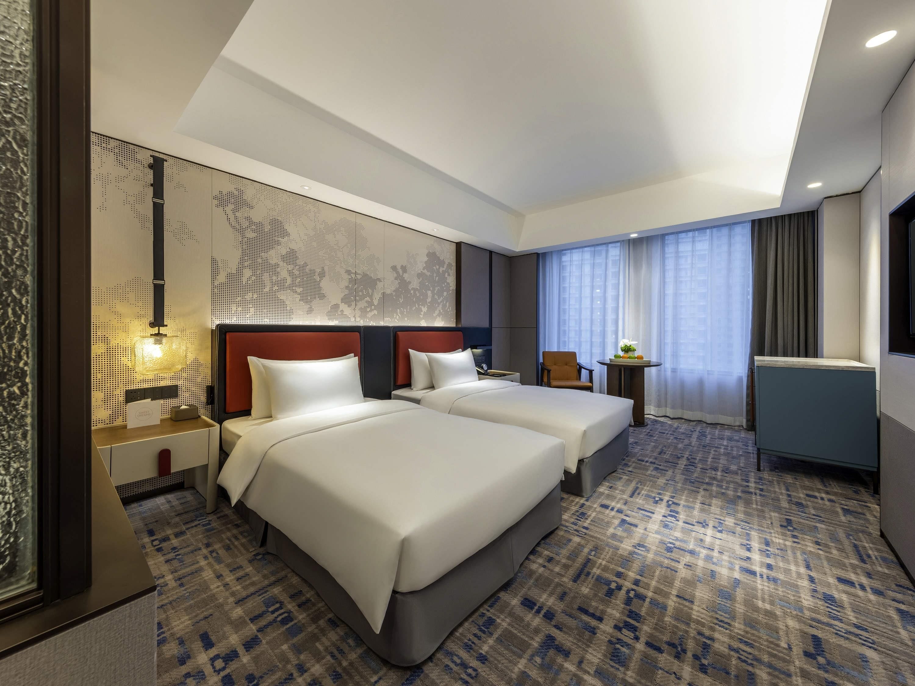 A hotel room with two beds, a large window with curtains, a desk with a chair, and a decorative wall panel.