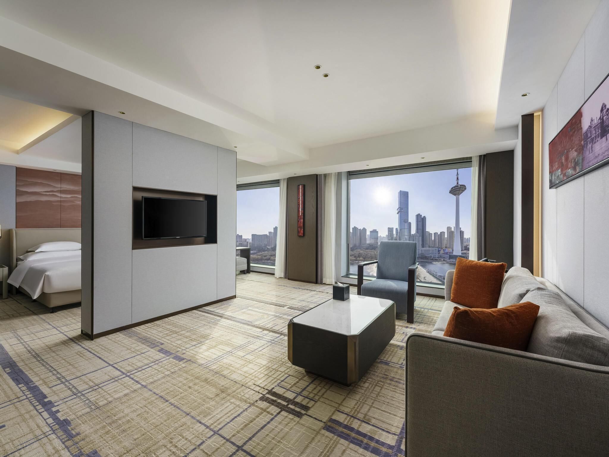 A hotel room with a bed, a sofa, a TV, a coffee table, and a large window offering a city view.