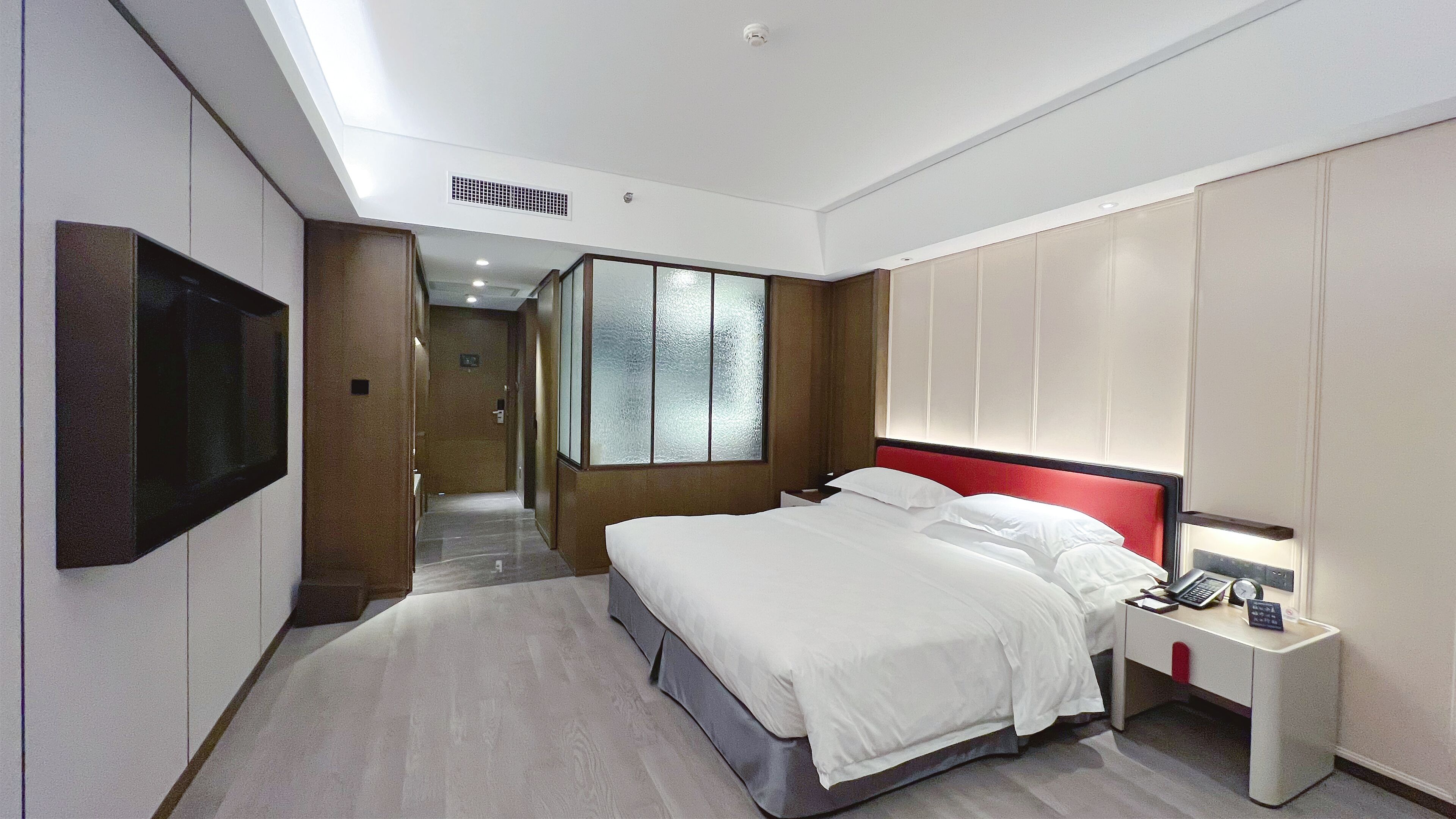 Et moderne hotelværelse med en stor seng, et sengebord med lampe, et fjernsyn ophængt på væggen og udsigt til gangen.