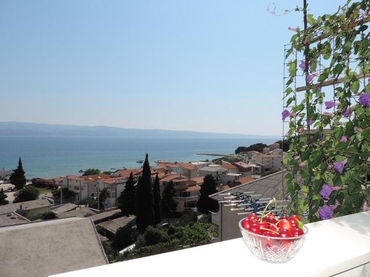 Un balcon donnant sur une ville côtière, une treille en métal ornée de fleurs violettes et un bol de cerises rouges.