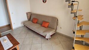 A living room with a wooden staircase, a sofa with orange cushions, and a small wooden table with a lace tablecloth.
