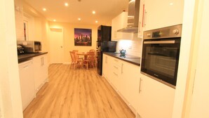 A compact kitchen with white cabinets, a black refrigerator, a microwave, and an oven. There is a dining table with chairs and a cityscape painting on the wall. - The Kyrenia (Weston-super-Mare)