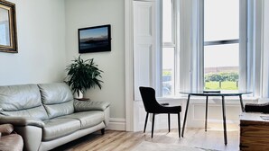 A living room with a leather sofa, a dining table with two chairs, a potted plant, and a large window with curtains. - The Kyrenia (Weston-super-Mare)