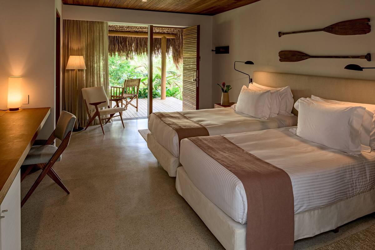 A hotel room with two beds, a desk, and a chair. There is a view of greenery outside.