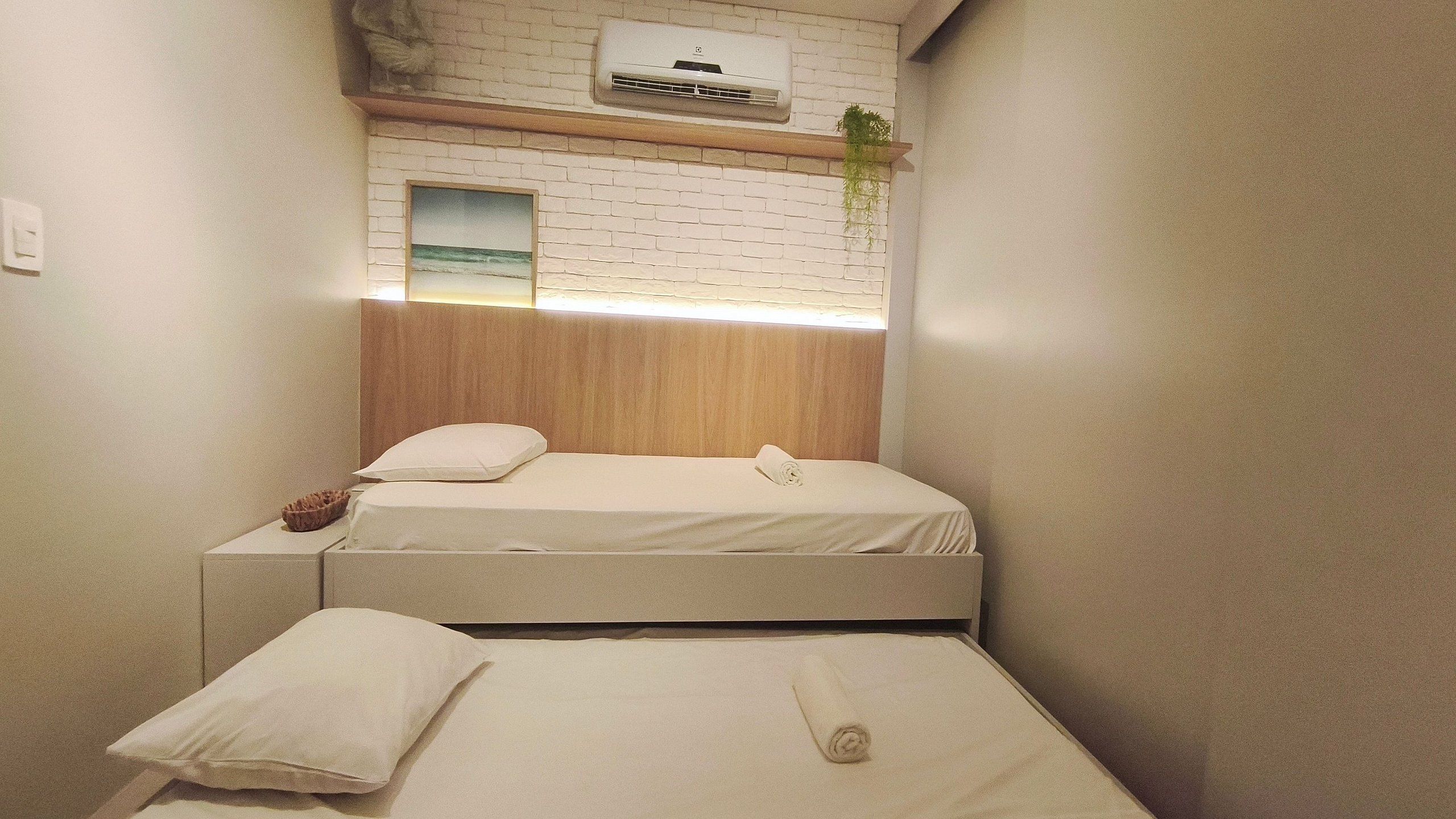 A small, clean room with a bed, a bedside table, a towel, a wall-mounted air conditioner, and a framed picture of a beach.