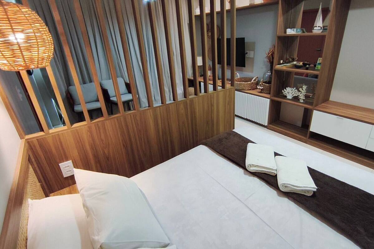Un dormitorio con una cama, un separador de madera, una estantería con objetos decorativos y una lámpara colgante.