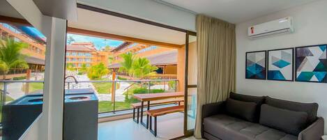 A modern hotel room with a balcony, a sofa, a dining table, and a view of the hotel complex.